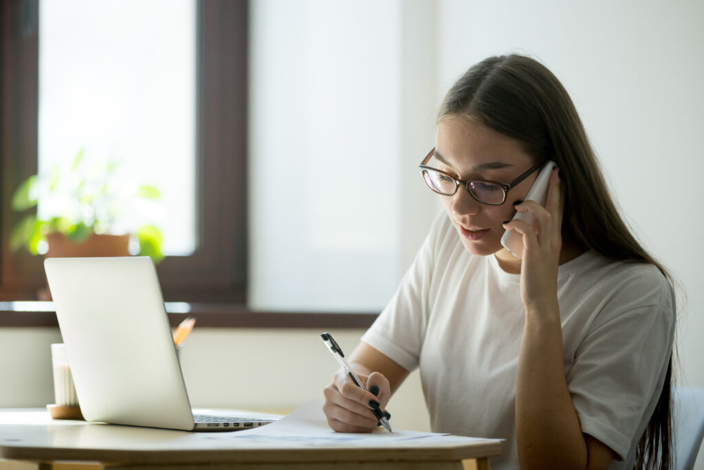A woman with glasses sits at a desk in a bright room, talking on the phone and taking notes, possibly setting an appointment for a mold inspection in the Bay Area.